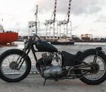 Classic motorcycle parked by a busy harbor with cranes and ships in the background.
